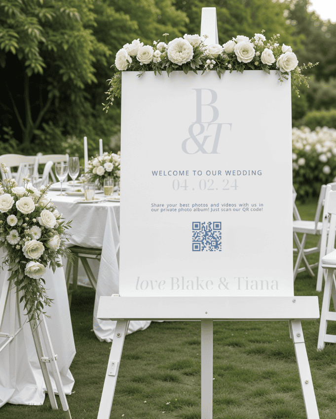 Hochzeits-Willkommensschild auf einer Staffelei mit cremefarbenen Blumen und Grünpflanzen, auf dem „Blake &amp; Tiana“, das Datum 04.02.24 sowie ein QR-Code für Fotos zu sehen sind.