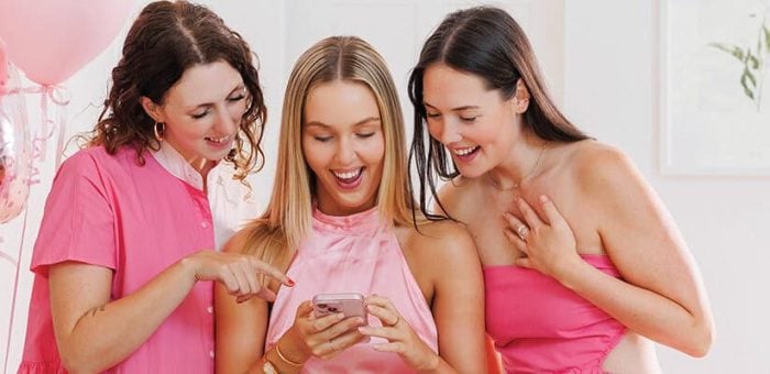 Three women in pink dresses gather around a phone, smiling and engaged. Pink balloons and flowers enhance the joyful, party atmosphere.