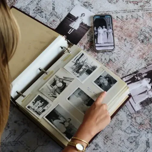 A woman reminisces on a vintage photo album on a patterned rug, surrounded by loose black-and-white photos and a smartphone displaying a GUESTPIX memorial gallery