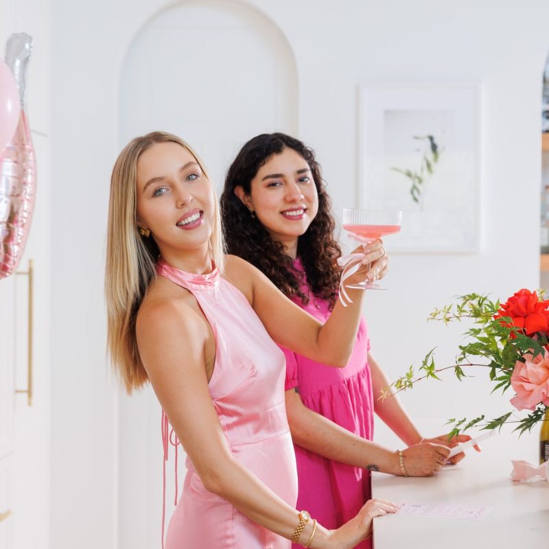 Dos mujeres celebrando en una fiesta: una lleva un vestido de satén rosa y sostiene un cóctel rosa, y la otra lleva un vestido rosa, con adornos florales y globos festivos en un entorno luminoso y elegante, que realza los momentos de alegría para compartir las fotos del evento con GUESTPIX.