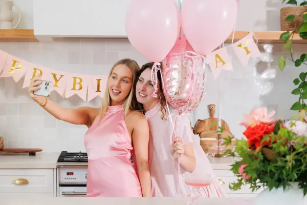Two women joyfully take a selfie in a kitchen. They wear pink dresses and hold pink balloons. A "Happy Birthday" banner and vibrant flowers decorate the scene.
