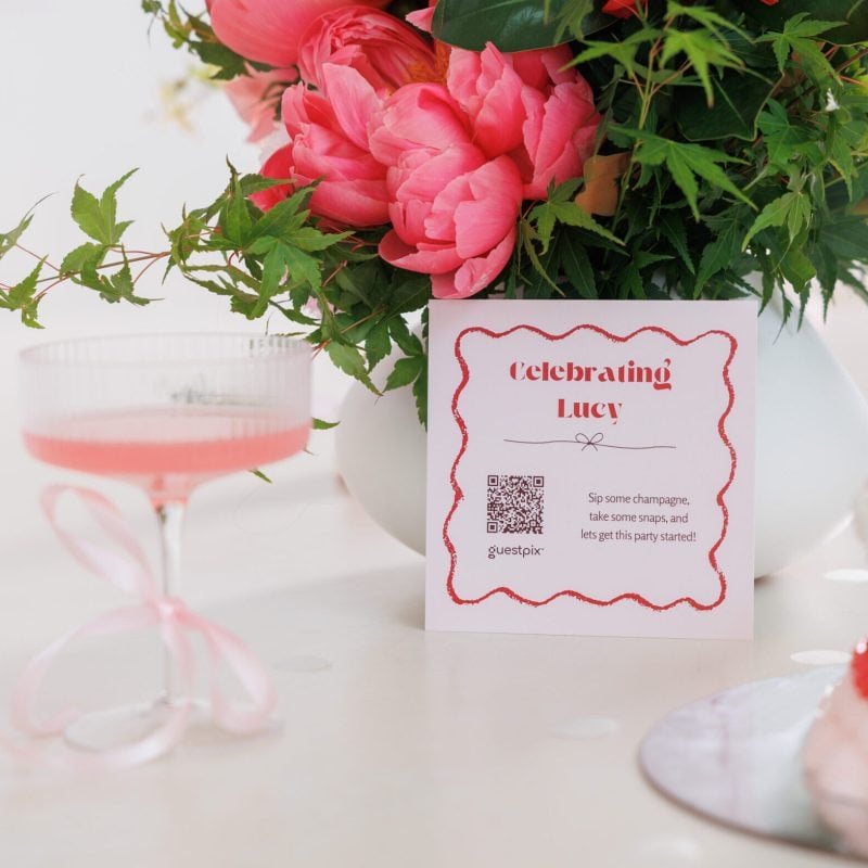 A vibrant bouquet of pink and red roses in a white vase with a card with a QR code on it, reading "Celebrating Lucy." And a pink cocktail in a glass with a bow.