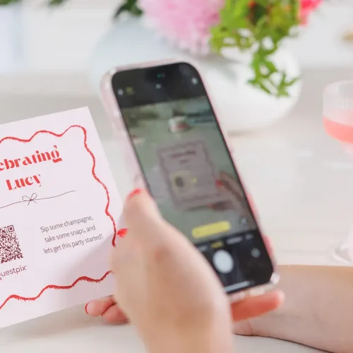 A person scans a GUESTPIX QR code on a pink invitation card with a phone. The card reads "Celebrating Lucy" in red. A pink cocktail and flowers are in the background.