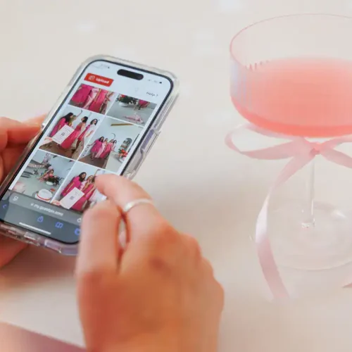 Person using a smartphone to view event photos on GUESTPIX platform, with a pink decorative glass in the background, highlighting easy photo sharing at events.