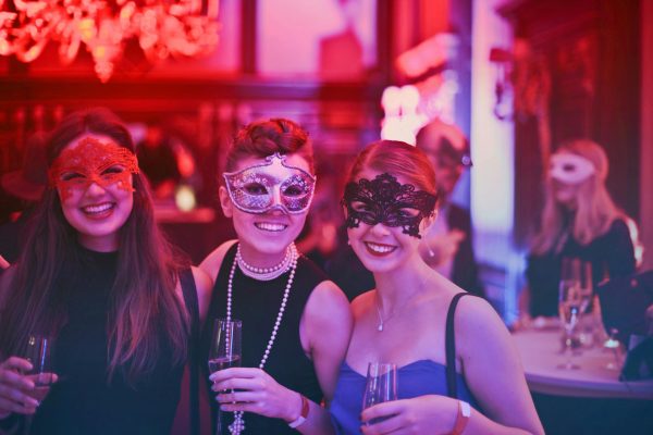 Elegant women wearing decorative masquerade masks enjoying a celebration at an event, holding champagne glasses in a lively, colourful party atmosphere.