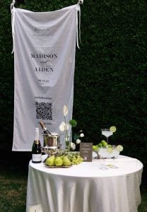 Bannière de mariage sur laquelle figurent les prénoms des mariés, Madison et Aiden, ainsi qu'un code QR permettant aux invités de partager leurs photos, à côté d'une table garnie de champagne, de pommes vertes et d'éléments décoratifs lors d'un mariage en plein air.
