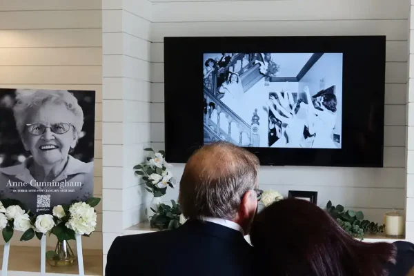 Man and woman embrace in front of a memorial. To the left, a portrait of an elderly lady. Black-and-white old photographs on the monitor showing memories in a slideshow.