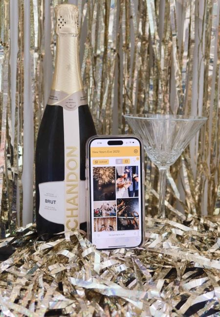A champagne bottle, smartphone displaying event photos, and a cocktail glass on a festive gold backdrop, highlighting the use of GUESTPIX for instant photo sharing at celebrations.