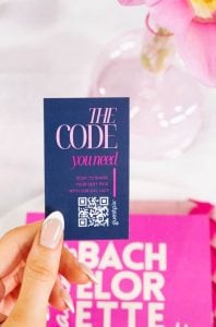 Gros plan sur une main tenant une carte avec un code QR permettant de partager des photos de l'événement à la « Bachelorette Suite », avec des fleurs roses et une pancarte rose en arrière-plan.
