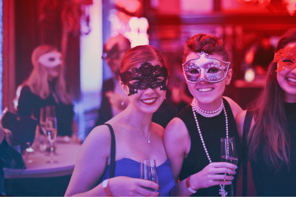 Guestpix event photo sharing, women in elegant masks enjoying drinks at masquerade party, colourful lighting, socialising at celebration, festive event image.