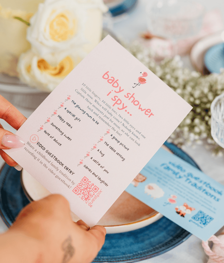Primo piano di un biglietto d'invito rosa per un baby shower dal titolo "I Spy", tenuto tra le mani, con l'illustrazione di un fenicottero, un elenco di indizi e un codice QR su una tavola imbandita.