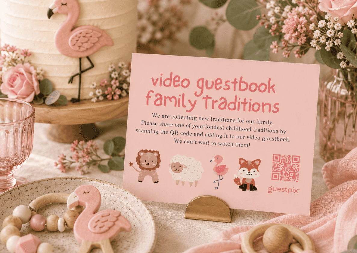 Cartello rosa con la scritta “video guestbook tradizioni di famiglia” su un tavolo dei dolci con un decoratore per torte a forma di fenicottero e decorazioni dai toni pastello su un tavolo fiorito.