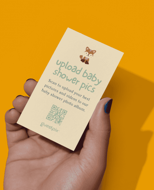 Una mano che regge un biglietto beige con la scritta “carica le foto del baby shower”, un piccolo disegno di una volpe e un codice QR, su uno sfondo giallo brillante.