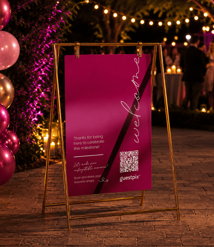 Cartello celebrativo in magenta su un cavalletto dorato con un codice QR per Guestpix, collocato durante una festa serale all’aperto con palloncini e lucine decorative.