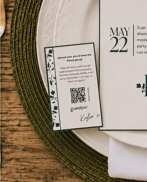 Carte d'invitation posée sur une assiette de présentation décorative, comportant un code QR et la mention « 22 MAI », ornée d'une bordure florale et d'une signature manuscrite « Kylie », disposée sur une table en bois avec une serviette blanche à proximité.