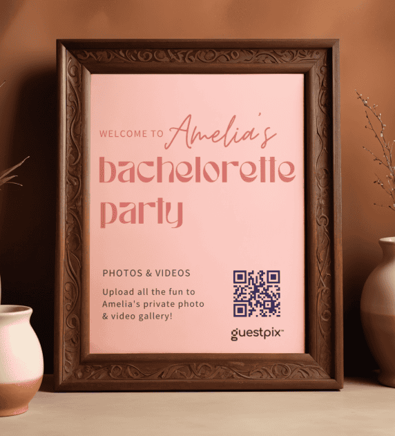 Invito incorniciato per un addio al nubilato, con la scritta "Benvenuti all'addio al nubilato di Amelia", un codice QR e il logo "guestpix", appoggiato su un tavolo con dei vasi nelle vicinanze.