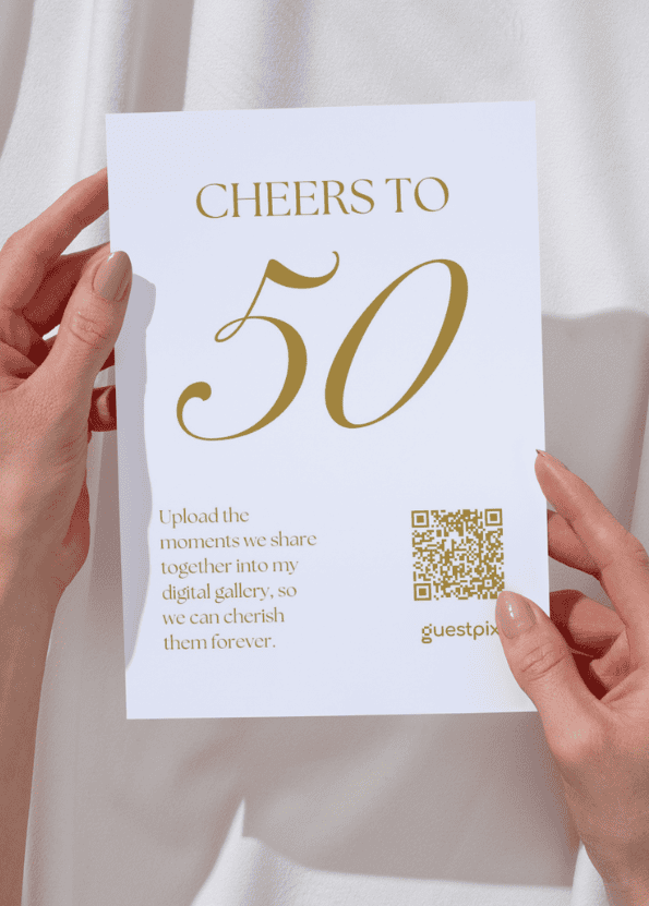 White card reading 'CHEERS TO 50' in gold, with a QR code and small gold text, held by two hands against a light background.