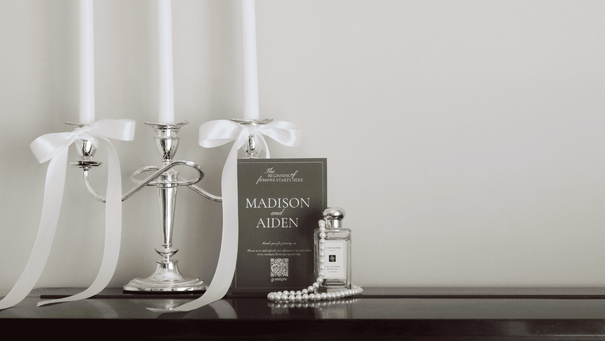 Decoración de mesa de boda con un candelabro plateado, cintas blancas y una tarjeta de invitación en la que se lee «MADISON y AIDEN», junto a un frasco de perfume y un collar de perlas sobre una superficie oscura.