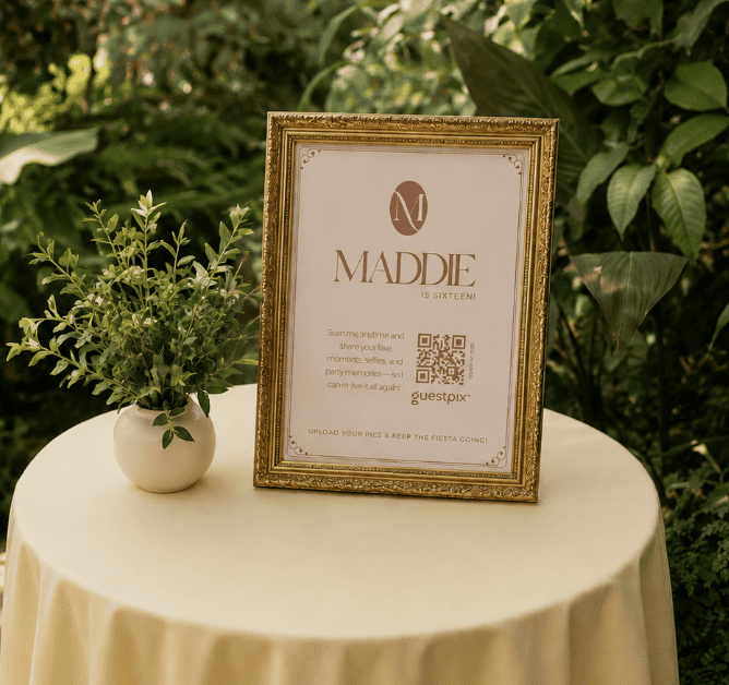 Un cartel dorado enmarcado en el que se lee «¡Maddie cumple dieciséis!», colocado sobre una mesa de color crema, junto a una pequeña planta en maceta, en un frondoso jardín al aire libre.
