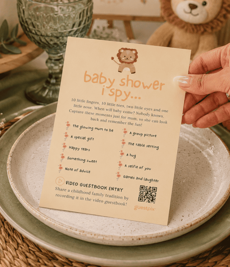 Carte « Je vois » pour une baby shower, tenue par une main, posée sur une assiette avec un ours en peluche en arrière-plan.