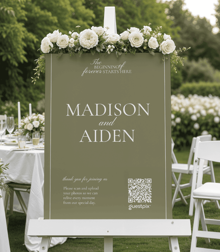 Cartello per matrimoni all'aperto con rose bianche, su cui sono riportati i nomi "MADISON e AIDEN" e un codice QR, collocato tra i tavoli e le sedie del ricevimento in giardino, circondato dal verde.