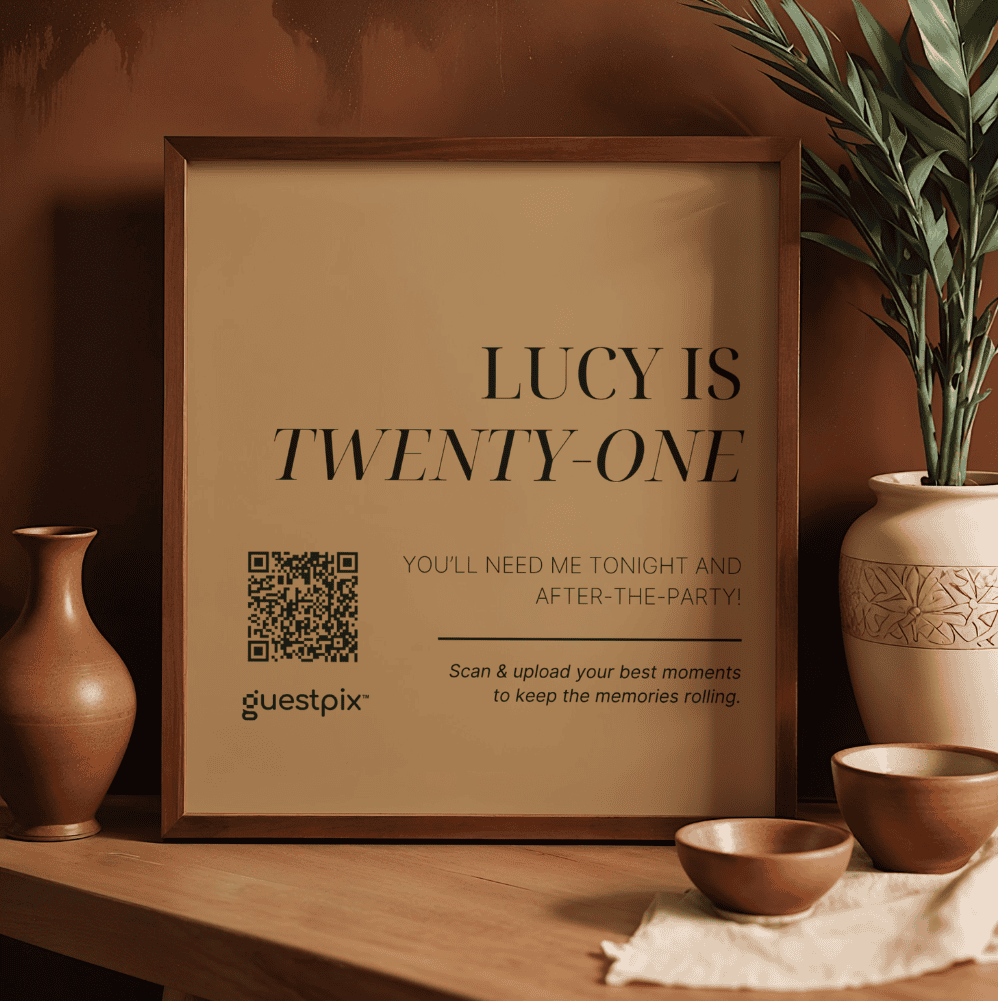 Poster incorniciato appoggiato su un tavolo di legno con la scritta “LUCY IS TWENTY-ONE”, accanto al quale sono visibili un codice QR e il logo di Guestpix.
