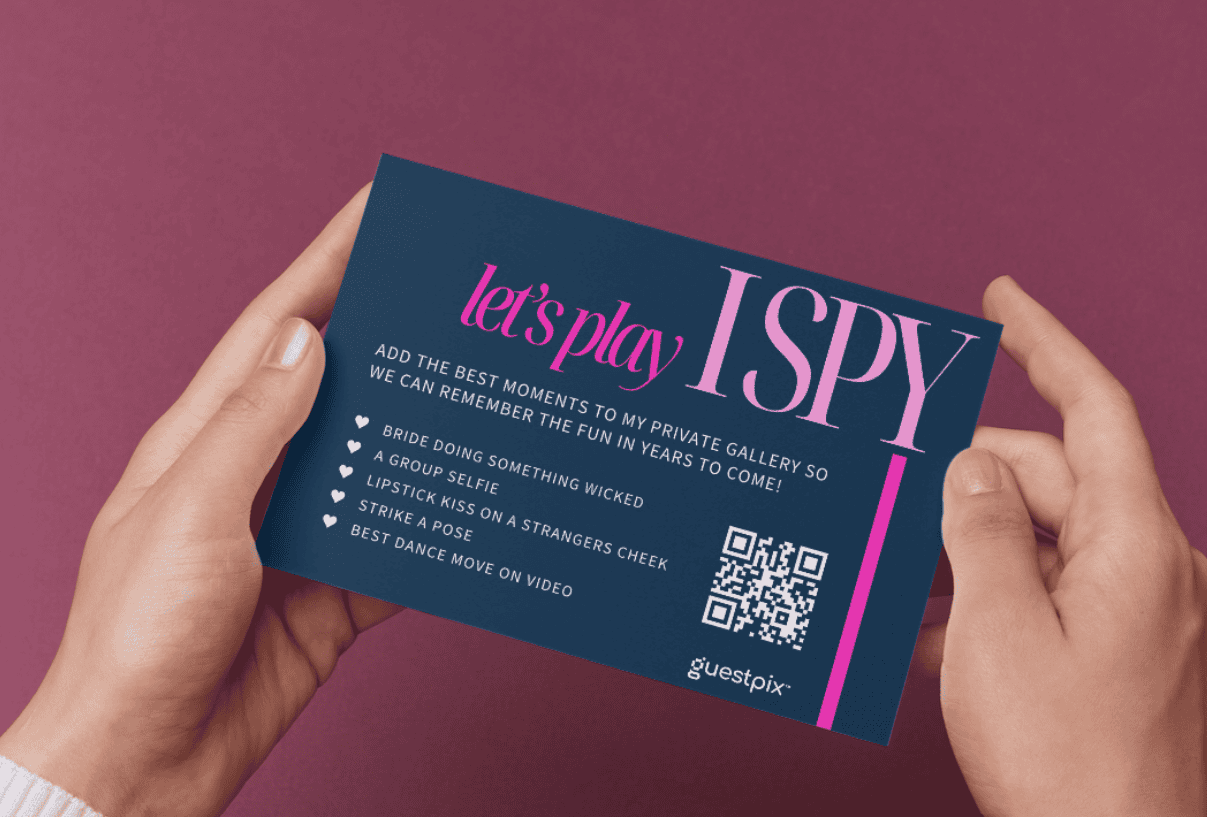 Le mani reggono un cartoncino blu scuro con la scritta "Giochiamo a ISPY", una striscia verticale rosa e un codice QR sul lato destro.