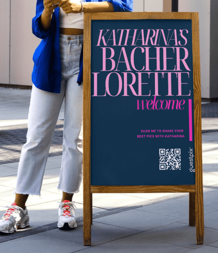 Una persona con una giacca blu e jeans chiari è in piedi accanto a un cartello con cornice in legno e pannello blu scuro su cui è scritto in lettere rosa “KATHARINA'S BACHER LORETTE welcome”; in basso a destra del cartello è visibile un codice QR.