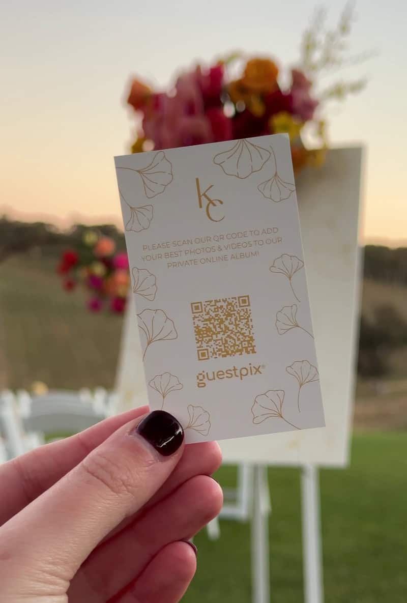 Primer plano de una tarjeta de la suite nupcial que incluye un código QR para que los invitados puedan acceder a las fotos del evento, con un fondo floral difuminado y un escenario al aire libre al fondo.