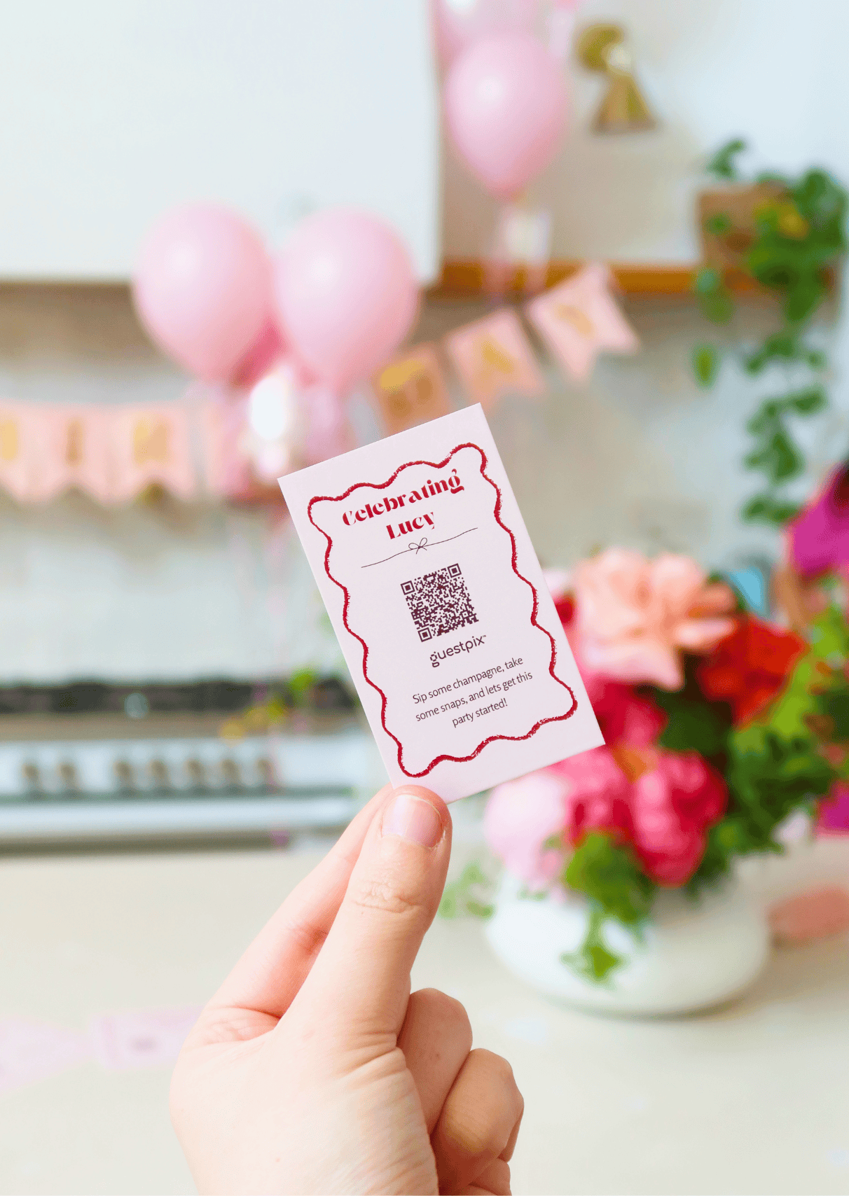 Card reading 'Celebrating Lucy' with a QR code, held up in front of pink balloons and floral decor in the background.