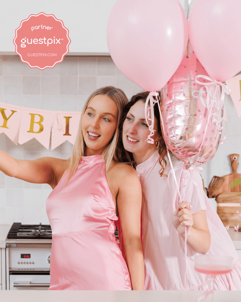 Zwei Frauen in rosa Outfits posieren für eine Geburtstagsfeier in einer Küche, im Hintergrund sind rosa Luftballons und ein Banner zu sehen.
