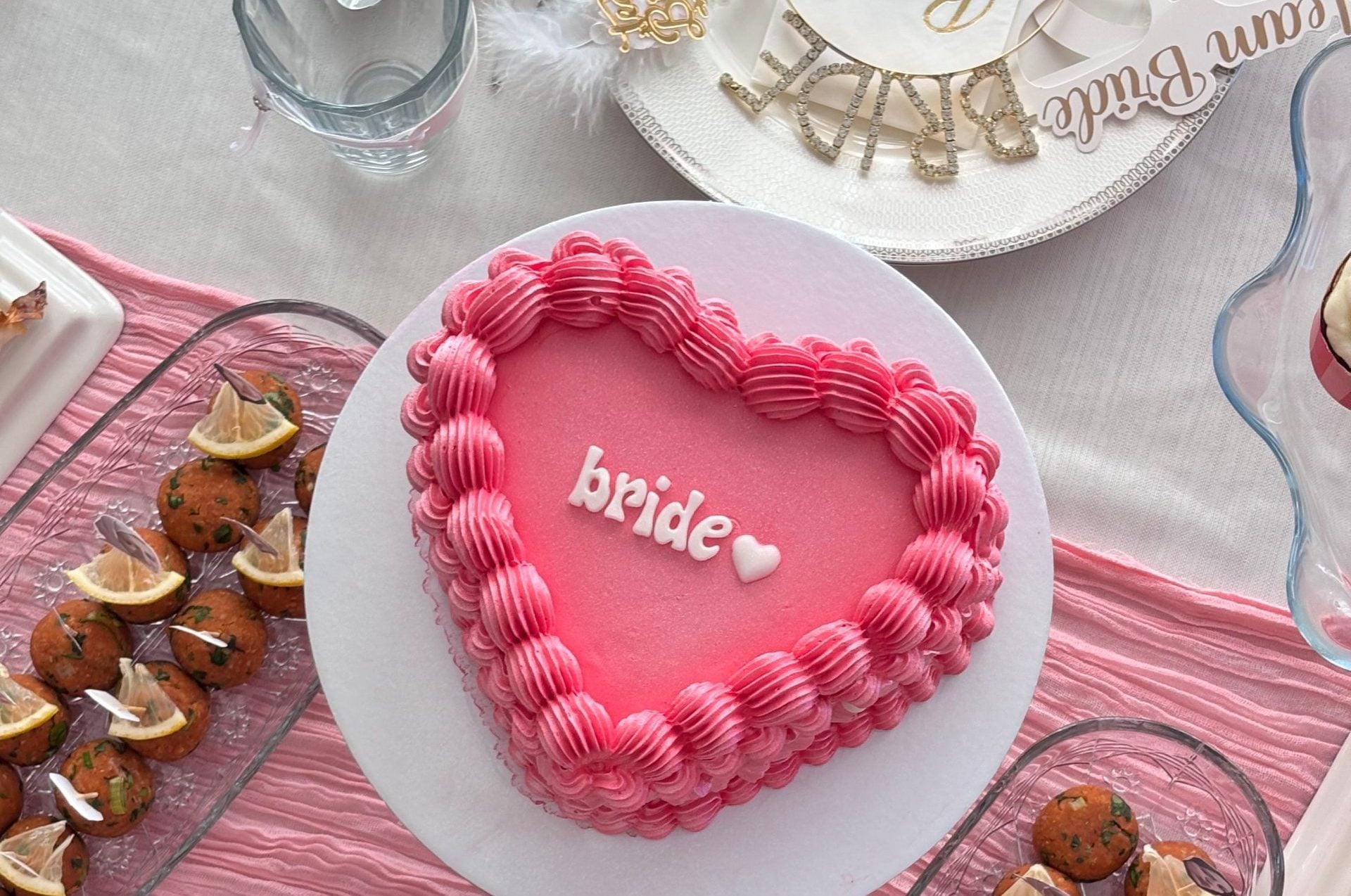 Pink heart-shaped wedding cake with a ruffled pink border and 'bride' in white icing on a white stand, centerpiece of the spread.