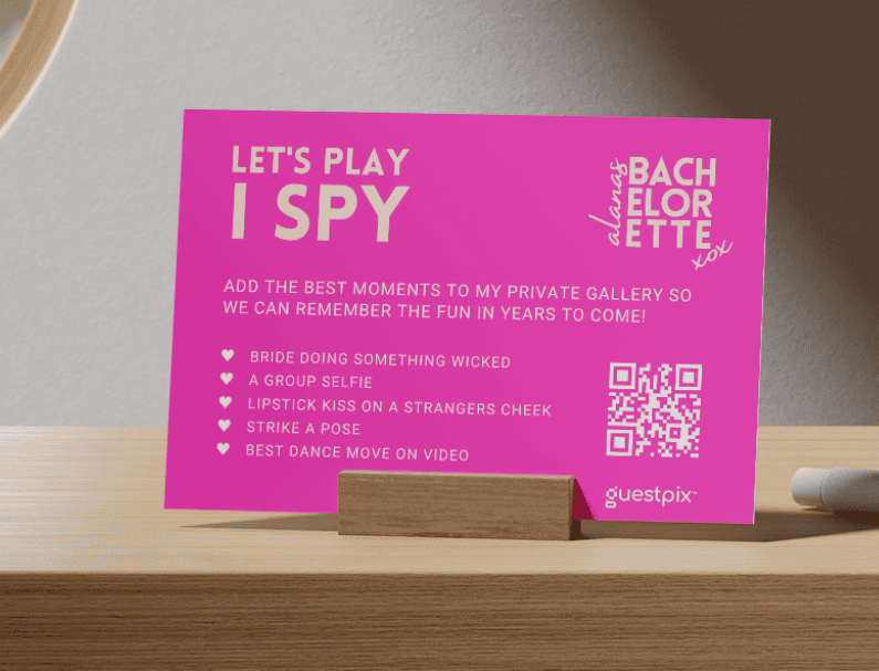 Tarjeta de color rosa brillante con el texto «Let's Play I Spy» sobre una mesa de madera, apoyada en un pequeño soporte de madera, con un código QR y el logotipo de Guestpix visibles en el lado derecho.