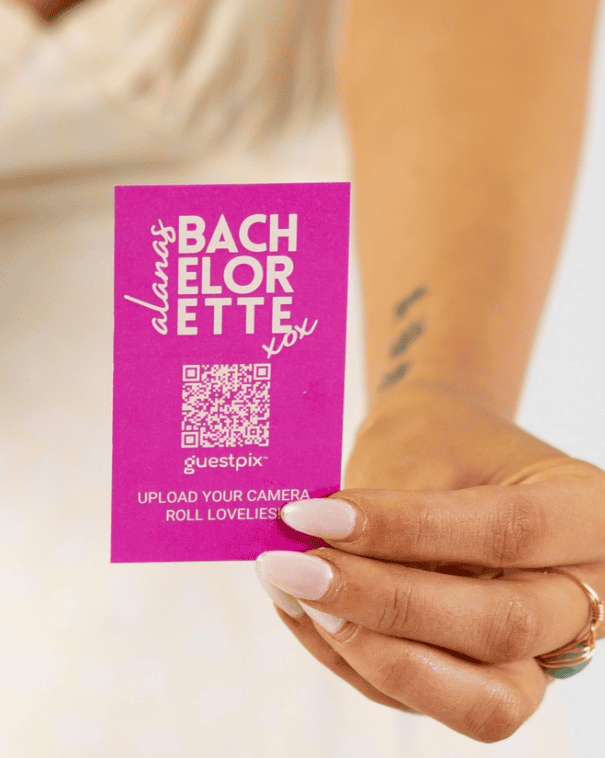 Tarjeta promocional morada de un servicio de fotos para despedidas de soltera con un código QR, sostenida por una mano, y el texto «SUBID LAS FOTOS DE VUESTRO ROLLO DE CÁMARA, QUERIDAS».
