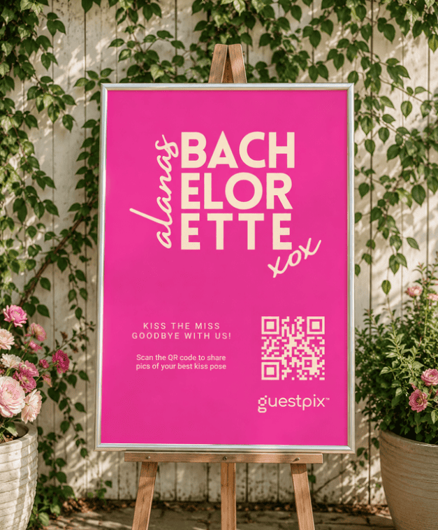 Cartel de color rosa brillante con el mensaje «Despedida de soltera» colocado en un caballete y con un código QR, en el que se invita a los invitados a compartir fotos en las que posan besándose con un fondo floral.