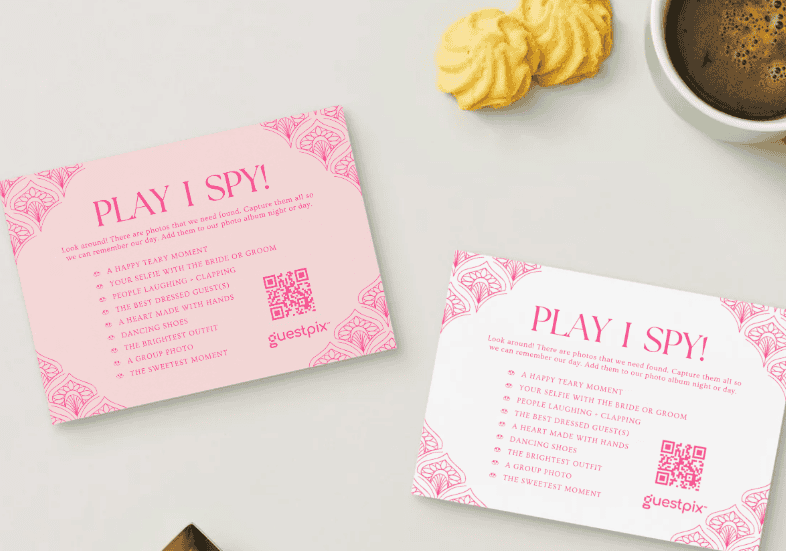 Deux cartes roses « PLAY I SPY » ornées de bordures décoratives et d'un code QR, posées sur une table claire, à côté de biscuits et d'une tasse de café.