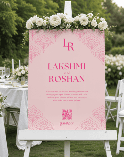 Hochzeitsschild auf einer weißen Staffelei mit einer rosa Tafel, auf der „Lakshmi und Roshan“ steht, und einer Blumengirlande darüber.