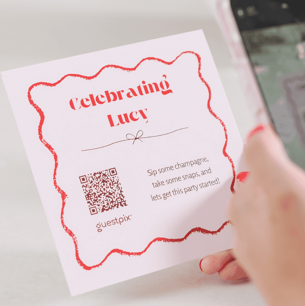 White invitation card with a red decorative border reading 'Celebrating Lucy' held by a hand.