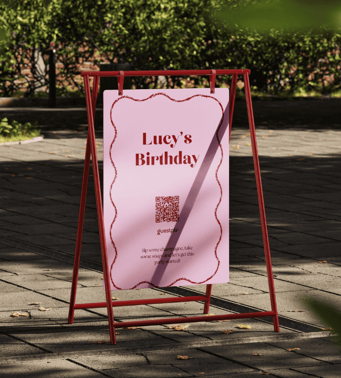 Cartel rosa de cumpleaños en el que se lee «Lucy's Birthday», colocado sobre un soporte rojo en forma de A en la acera, con un código QR y un borde festoneado decorativo.