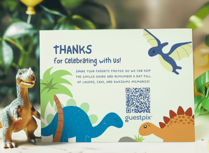 Eine Dankeskarte mit Dinosaurier-Motiv und der Aufschrift „Danke, dass ihr mit uns gefeiert habt!“ sowie einem QR-Code; darauf sind eine blaue, stegosaurierähnliche Figur und weitere Dinosaurier zu sehen, daneben steht eine Dinosaurier-Spielzeugfigur auf einem Tisch.