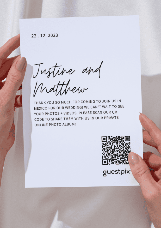 Eine mit beiden Händen gehaltene Hochzeitseinladungskarte, auf der die Namen Justine und Matthew sowie ein QR-Code zum Teilen von Fotos über Guestpix zu sehen sind.
