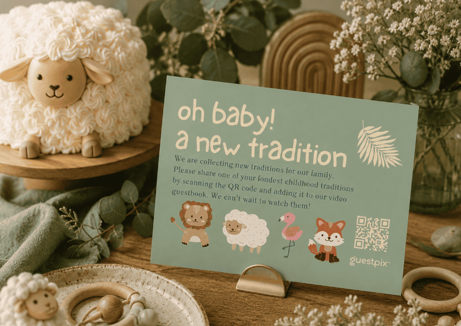 Auf einer mintfarbenen Karte, die auf einem Holztisch liegt, steht „Oh Baby! Eine neue Tradition“ – dazu gibt es einen QR-Code und niedliche Tierillustrationen; ein Plüschschaf und Grünpflanzen umgeben die Karte.