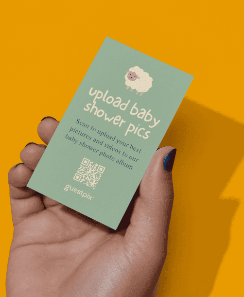 Eine mintgrüne Karte, die von einer Hand vor einem orangefarbenen Hintergrund gehalten wird; der Text lautet „Baby-Shower-Fotos hochladen“ und zeigt ein kleines Schaf, einen QR-Code sowie eine Anleitung zum Hochladen von Fotos in ein Baby-Shower-Album.