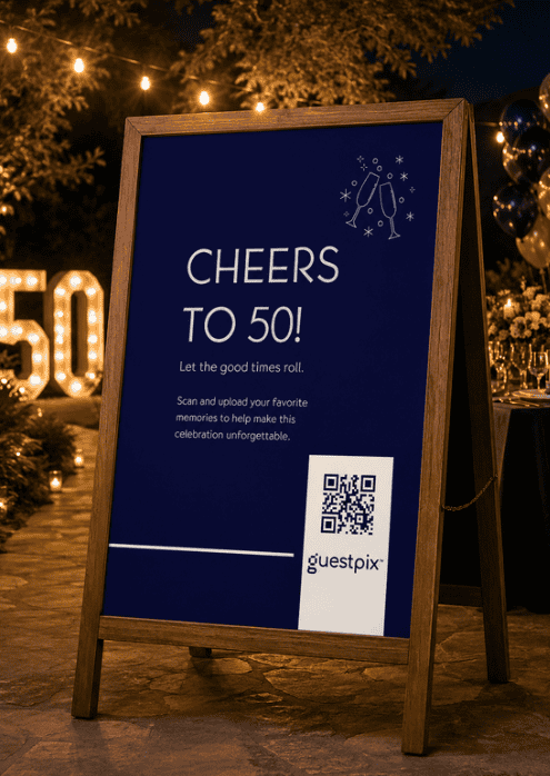 Un cartello blu per l'anniversario, montato su una cornice di legno, recita "CHEERS TO 50!" ed è provvisto di un codice QR per Guestpix in occasione di una festa all'aperto.
