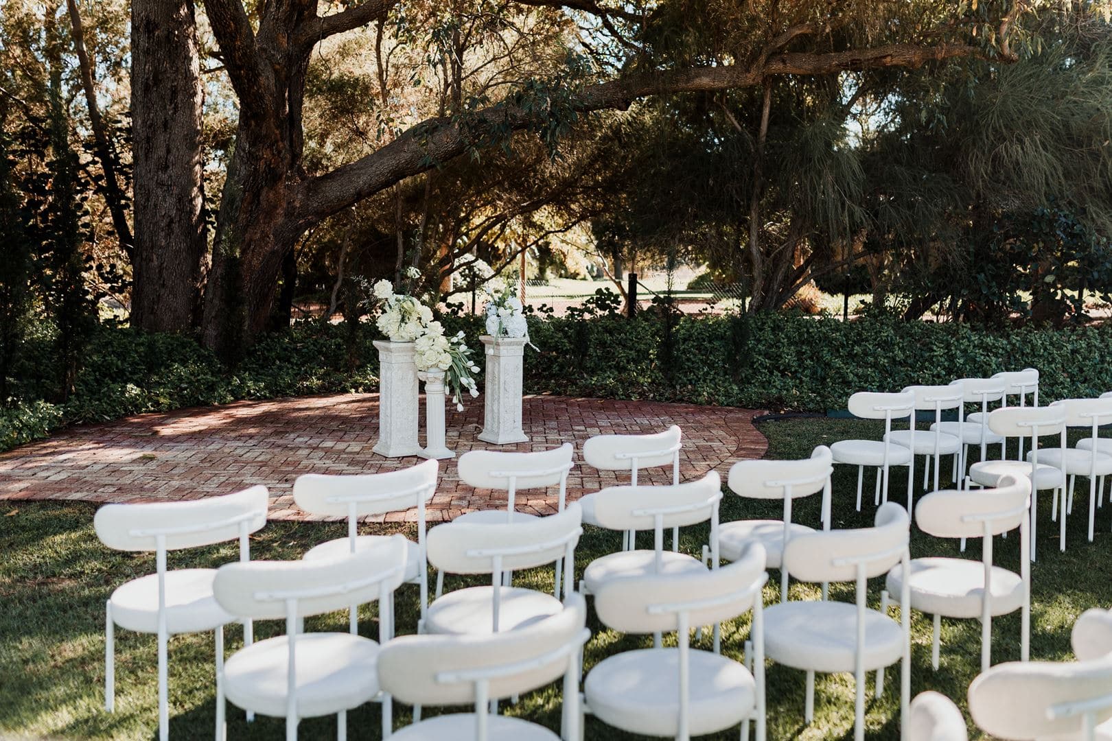 Wedding ceremony setup in a lush outdoor garden with white chairs and floral arrangements under large trees. - GUESTPIX | #1 QR Code Event Photo Sharing Platform Aménagement d'un lieu de mariage en plein air